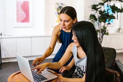 Two women in front of a laptop while they point at the screen and one is explaining something to the